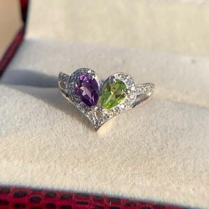 May include: A silver ring featuring a heart-shaped design with two pear-cut gemstones. One gemstone is a vibrant purple, the other is a bright green. The ring is encrusted with small, clear accent stones.