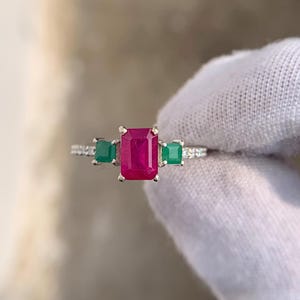May include: A silver ring with a central, rectangular, deep red gemstone, and two square, green gemstones either side. The ring band is set with small, clear accent stones, creating a luxurious and eye-catching design.