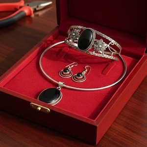 May include: A silver jewelry set with black oval stones, including a necklace, bracelet, and earrings, displayed in a red velvet-lined wooden box. The bracelet features floral designs. Pliers are visible in the background.