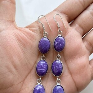 Puede incluir: Pendientes colgantes con piedras preciosas de charoíta morada ovaladas engastadas en metal plateado. Cada pendiente tiene tres piedras, cada una con un patrón arremolinado único. El texto "CHAROITE GEMSTONE" es visible en la parte inferior.