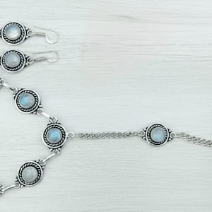 Può includere: Un set di gioielli color argento composto da un braccialetto, un paio di orecchini e un anello. Ogni pezzo presenta un design circolare con una pietra azzurra al centro, circondata da un bordo ornato. Il braccialetto è collegato da una catena.