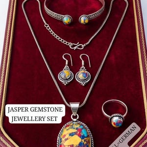 May include: A complete jasper gemstone jewelry set, including a necklace, bracelet, earrings, and ring, displayed in a red jewelry box. The set features silver-toned metal and colorful, patterned gemstones. The text "JASPER GEMSTONE JEWELLERY SET" is visible.