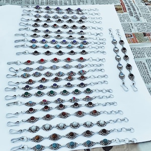 Op de afbeelding: Een verzameling zilverkleurige armbanden met verschillende edelsteenaccenten, waaronder rode, blauwe, groene en witte stenen. De armbanden zijn gerangschikt op een wit oppervlak, waarbij hun ingewikkelde ontwerpen en diverse kleuren worden getoond.