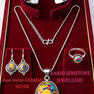 Op de afbeelding: Een zilveren sieradenset bestaande uit een manchetarmband, ketting, oorbellen en ring, elk met een kleurrijke edelsteen. De set wordt gepresenteerd in een rode fluwelen doos. De tekst "RAKHI JEMSTONE JEWELLERS" is zichtbaar.