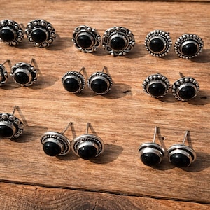 May include: A selection of silver stud earrings with black onyx stones. The earrings have different designs, including floral and circular patterns. The earrings are presented on a wooden surface.