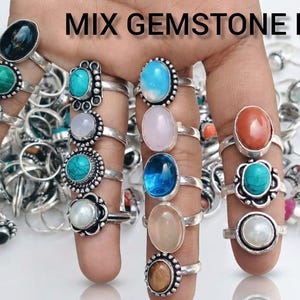 May include: A collection of gemstone rings with various colored stones, including blue, green, pink, and orange, displayed on fingers. The rings have silver bands and are set with oval and round stones. The text "MIX GEMSTONE RING" is at the top.