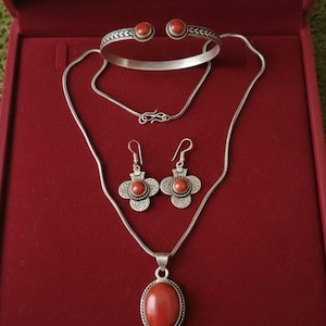 Può includere: Un set di gioielli composto da un bracciale rigido in argento, un paio di orecchini pendenti e una collana con ciondolo, tutti caratterizzati da pietre lisce di colore rosso-arancio incastonate in argento con dettagli a corda e filigrana.