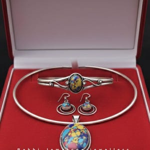 Könnte beinhalten: Ein Schmuckset in einer roten Box, bestehend aus einem Armband, Ohrringen und einem Anhänger. Der Schmuck weist farbenfrohe, ovale Edelsteine auf. Der Markenname "Rakhi Jemstone jewellers" ist am unteren Rand der Box aufgedruckt.