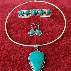 May include: A silver jewelry set featuring turquoise stones. The set includes a necklace with a teardrop pendant, a cuff bracelet, and a pair of earrings. The jewelry is displayed on a red surface.