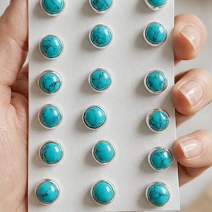 May include: A collection of turquoise gemstone stud earrings. Each round earring features a vibrant blue-green stone set in a silver-colored bezel. The earrings are displayed on a white card. Text at the bottom reads "TURQUOISE GEMSTONE STUD EARRING."