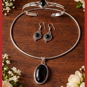 Può includere: Un set di gioielli in argento con una pietra nera. Il set include un braccialetto, una collana e un paio di orecchini. I gioielli sono esposti su una superficie di legno, con il testo "Rakhi Jemstone jewellers" in alto. Accenti floreali incorniciano i gioielli.