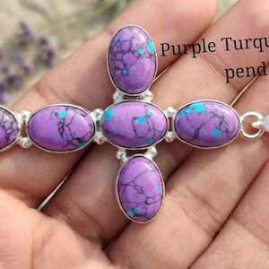 Peut inclure: Pendentif en argent en forme de croix orné de pierres ovales en turquoise violette veinées de bleu et de noir. Le texte "Purple Turquoise Cross pendant" est visible. Le pendentif est tenu dans une main.