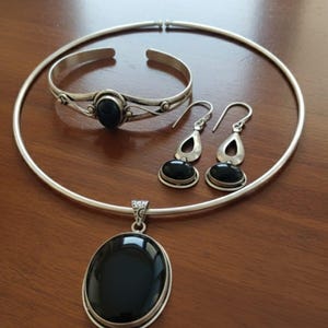 May include: A silver jewellery set comprising a necklace, bracelet, and earrings. The set includes a pendant with a large oval black stone, a matching bracelet, and drop earrings, all with similar black stones.