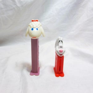 May include: Two vintage PEZ dispensers. One features a white sheep with a red bow and blue eyes on a purple base. The other is a white rabbit with a pink nose and a red base. Both are classic candy dispensers.
