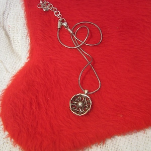 Puede incluir: Un collar plateado con un colgante circular con un dise&ntilde;o intrincado y arremolinado. El colgante cuelga de una delicada cadena, sobre una superficie roja vibrante y difusa. El collar es una joya.