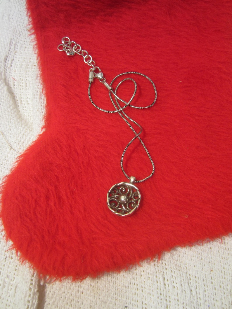 Puede incluir: Un collar plateado con un colgante circular con un dise&ntilde;o intrincado y arremolinado. El colgante cuelga de una delicada cadena, sobre una superficie roja vibrante y difusa. El collar es una joya.