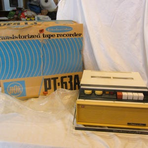Könnte beinhalten: Vintage beigefarbener und schwarzer Tonbandrekorder mit passender Box. Auf der Box steht "transistorized tape recorder" mit blauen Wellenmustern. Der Rekorder hat einen silbernen Griff und mehrere Knöpfe. Modellnummer PT-63A.
