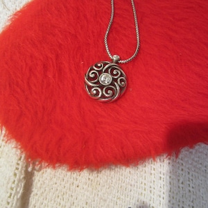 Puede incluir: Collar con colgante plateado con un dise&ntilde;o circular con patrones arremolinados y una gema transparente central. El colgante cuelga de una delicada cadena. El collar descansa sobre una superficie roja vibrante y esponjosa.