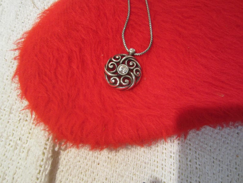 Puede incluir: Collar con colgante plateado con un dise&ntilde;o circular con patrones arremolinados y una gema transparente central. El colgante cuelga de una delicada cadena. El collar descansa sobre una superficie roja vibrante y esponjosa.