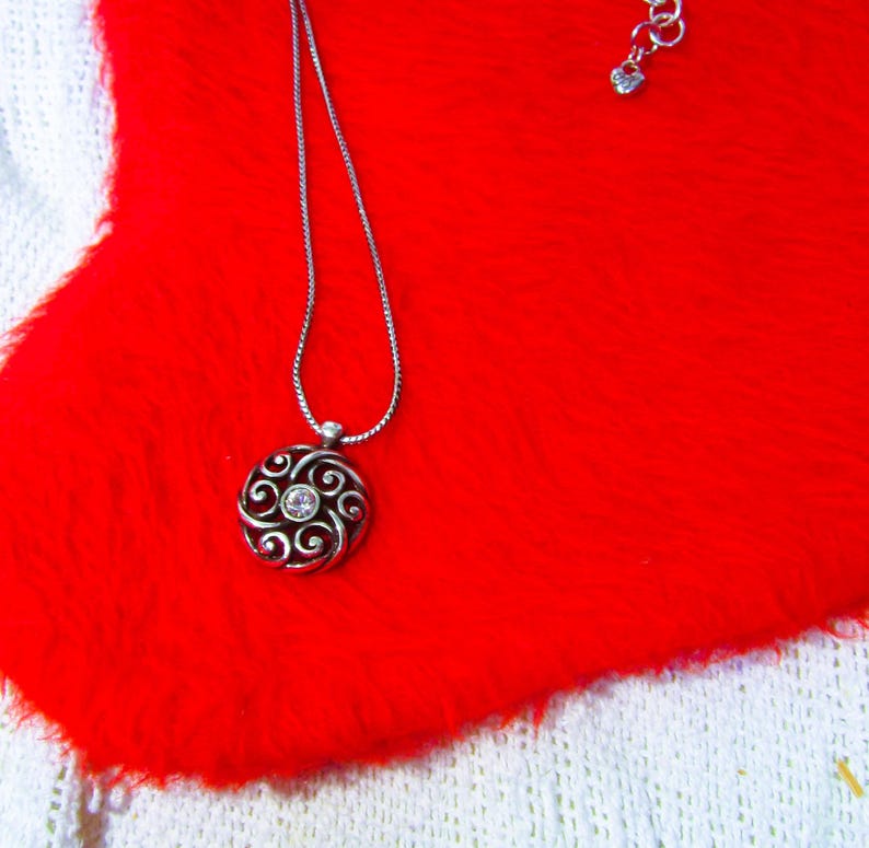 Puede incluir: Collar plateado con un colgante circular con un dise&ntilde;o en espiral y una piedra central transparente. El collar descansa sobre una superficie roja vibrante y texturizada, con una tela blanca tejida en el fondo.