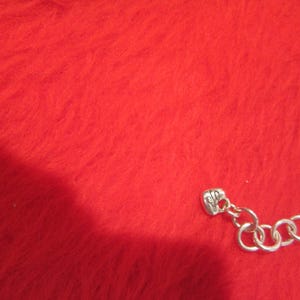 Puede incluir: Un collar plateado con un dise&ntilde;o de cadena de serpiente. El collar se muestra sobre una superficie roja texturizada. El collar tiene un peque&ntilde;o dije y una serie de anillos conectados en el cierre.