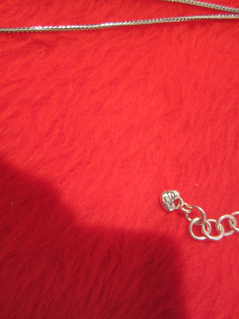 Puede incluir: Un collar plateado con un dise&ntilde;o de cadena de serpiente. El collar se muestra sobre una superficie roja texturizada. El collar tiene un peque&ntilde;o dije y una serie de anillos conectados en el cierre.