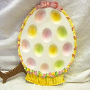 May include: An egg-shaped ceramic serving dish with 12 egg-shaped indentations. The dish is white with pastel-colored egg-shaped accents. It has a yellow basket-weave border with a pink bow at the top and a floral basket base.