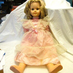 May include: A doll with blonde hair and blue eyes wearing a pink satin dress with a white tulle overlay featuring a ruffled collar and a pink ribbon bow at the waist. The doll has light skin, articulated limbs, and is seated in a forward-facing position.