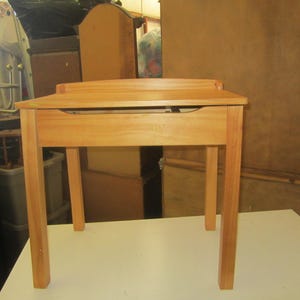 May include: A light brown wooden desk featuring a flat rectangular top surface, four straight legs, and a horizontal front panel. The desk has a smooth finish and a simple construction with a slightly raised back edge along the top surface.