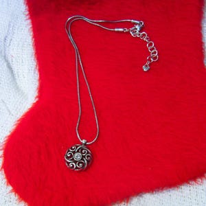 Puede incluir: Un collar plateado con un colgante circular con un dise&ntilde;o en espiral y una piedra transparente central. El collar se muestra sobre una superficie roja y difusa, con un fondo blanco tejido.