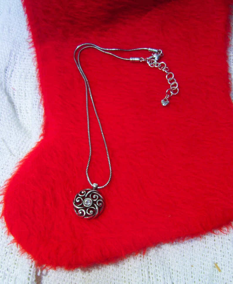 Puede incluir: Un collar plateado con un colgante circular con un dise&ntilde;o en espiral y una piedra transparente central. El collar se muestra sobre una superficie roja y difusa, con un fondo blanco tejido.