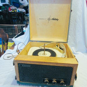 May include: Vintage Webcor Holiday record player with a tan and blue exterior. The open lid reveals the turntable and a black vinyl record. The front features a dark blue speaker cover and control knobs. The record player is a retro audio device.