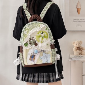 Könnte beinhalten: Ein grün-weiß karierter Rucksack mit einer transparenten Vordertasche, die mit weißer Spitze, einem grünen Apfelmotiv und kleinen Plüschverzierungen geschmückt ist. Der Rucksack hat braune Akzente, einen Tragegriff und Schultergurte mit weißen Bändern.