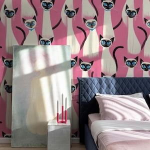 Pode incluir: Uma parede rosa com um padrão repetido de gatos siameses com olhos azuis e marcas pretas. Uma cama com cabeceira acolchoada azul e roupa de cama rosa está em primeiro plano. Uma pintura e uma mesa de cabeceira branca com um castiçal rosa também são visíveis.