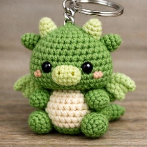 May include: A handmade green crochet dragon keychain with a cream-colored belly. The dragon has black eyes, small wings, and blush-colored cheeks. The keychain is attached to a silver metal ring.