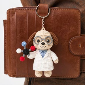 Puede incluir: Un llavero de perro de ganchillo con gafas y bata de laboratorio, sosteniendo un modelo molecular. El perro tiene orejas marrones y está unido a un llavero plateado. El fondo es una cartera de cuero marrón.