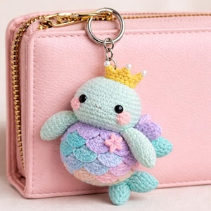 May include: A crochet mermaid turtle keyring. The turtle has a light blue head, a colourful tail with pastel scales, and a gold crown. The keyring is attached to a pink wallet with a gold zip.