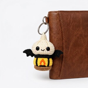 May include: A handmade crochet keychain featuring a smiling, light-colored creature with black bat wings and a yellow and black lantern body. The keychain is attached to a silver ring, next to a brown leather wallet.