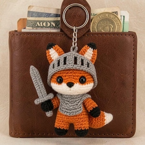 Puede incluir: Llavero de zorro de ganchillo, naranja, blanco y negro, diseñado como un caballero con casco y espada grises. El zorro está unido a una cartera de cuero marrón, con un llavero de metal.