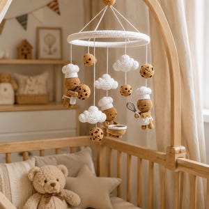 May include: A crochet baby mobile featuring tan gingerbread figures wearing white chef hats and aprons, white clouds, and brown-spotted cookies suspended from a white circular frame on a wooden arm. The figures hold kitchen tools.