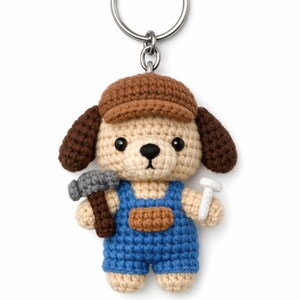 May include: A crocheted dog keyring wearing a brown cap and blue dungarees, holding a hammer and a nail. The dog has brown ears and a light beige body. The keyring is attached to a silver metal ring.