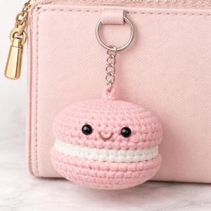 May include: A pink crocheted macaron keychain with a smiling face, black eyes, and a white filling. The keychain is attached to a silver chain and ring, and is next to a pink wallet.