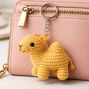 Puede incluir: Un llavero de camello tejido a ganchillo en amarillo, con una cara sonriente y ojos negros. El amuleto está unido a un llavero plateado, sobre un fondo de cartera de cuero rosa. Un accesorio único y encantador.