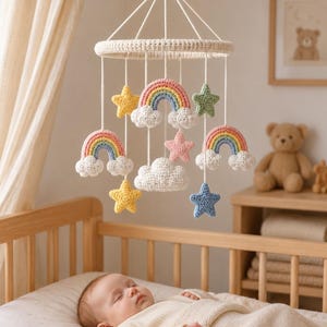 May include: A hanging nursery mobile featuring crocheted elements suspended from a circular frame. The mobile includes three colourful rainbows with white clouds, three stars in yellow, pink, and blue, one green star, and one white cloud.