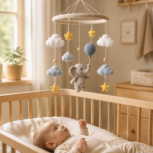 May include: A crochet baby mobile featuring a central grey elephant holding a blue balloon, surrounded by white and light blue clouds and yellow stars suspended from a circular frame. The elements are crafted with yarn and wooden beads.
