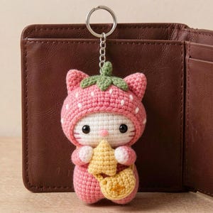Op de afbeelding: Een gehaakte sleutelhanger van een wit katfiguur met een roze aardbeienkapje met groene blaadjes. De kat heeft zwarte ogen, een roze neusje, houdt een klein geel stervormig object vast en draagt een bijpassend geel schoudertasje.