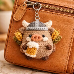 May include: A handmade crochet keychain depicting a pig in a Viking helmet, holding a beer mug. The pig is brown with cream horns and a pink snout. The keychain is attached to a brown leather wallet with a zip.