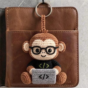 May include: Crocheted monkey keychain with glasses, a black shirt, and a gray sign with code symbols. The brown and beige monkey is attached to a silver keyring. The keychain is set against a brown leather wallet.
