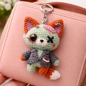 May include: A handmade crochet fox keychain with a zombie-like appearance. The fox has a green body, orange and pink ears, and a gray jacket. It features a black eye and an X over the other eye. The keychain is attached to a silver ring.