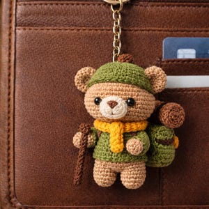Peut inclure: Porte-clés en forme d'ours crocheté, avec un chapeau, un pull et un sac à dos verts. L'ours porte une écharpe jaune et un bâton de marche marron. Le porte-clés est fixé à une chaîne dorée, sur fond de cuir marron.